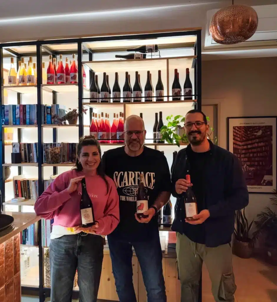 Drei Personen halten Weinflaschen von Markatselis vor einem Weinregal.