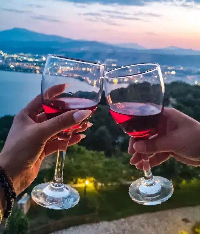 Δύο χέρια τσουγκρίζουν ποτήρια με κόκκινο κρασί, με θέα λίμνη και βουνά την ώρα του σούρουπου
