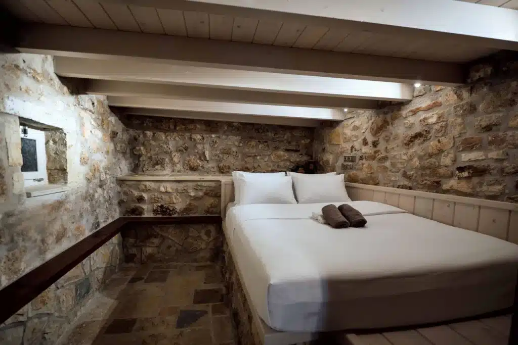 Suite bedroom with a double bed, dark wooden beams and a stone accent wall, Valaes Guesthouse, Foiniki