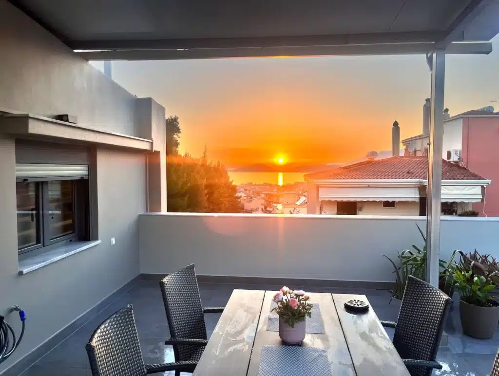 Covered balcony with a table and chairs facing the sunset over rooftops