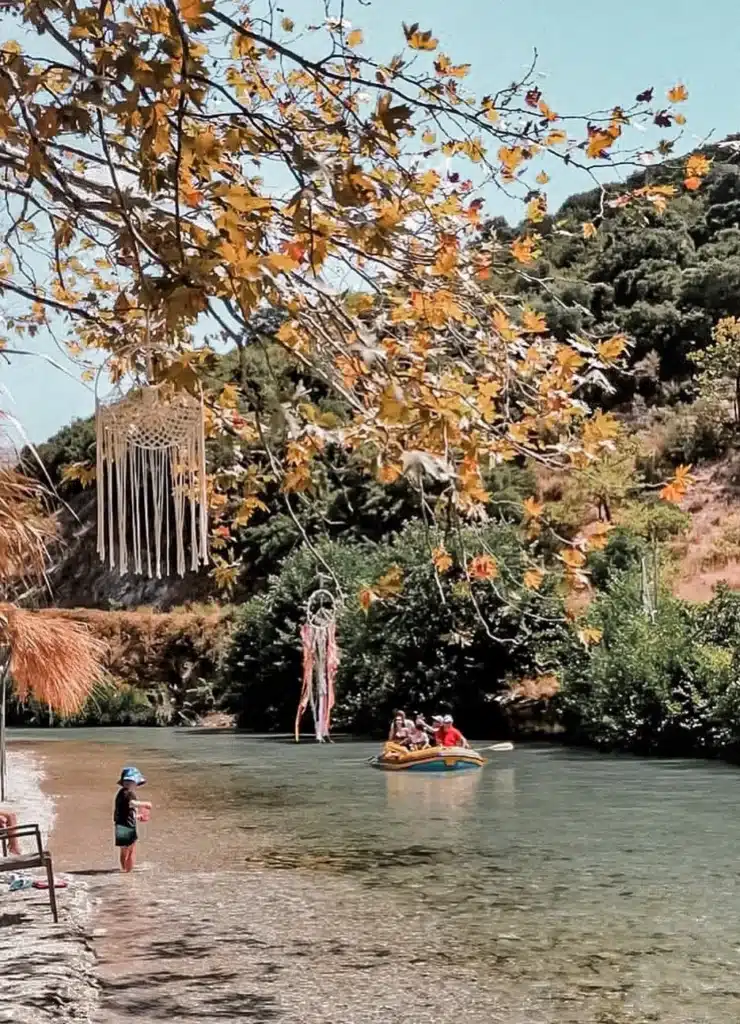 River bank shaded by plane trees with hanging macramé decorations, a child at the water’s edge and a small boat on the river