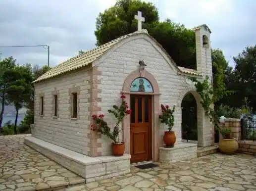 Το ξωκλήσι της Αγίας Παρασκευής στην Πλαταριά από το πλάι, με κεραμοσκεπή, σταυρό και γλάστρες, και τη θάλασσα στο βάθος